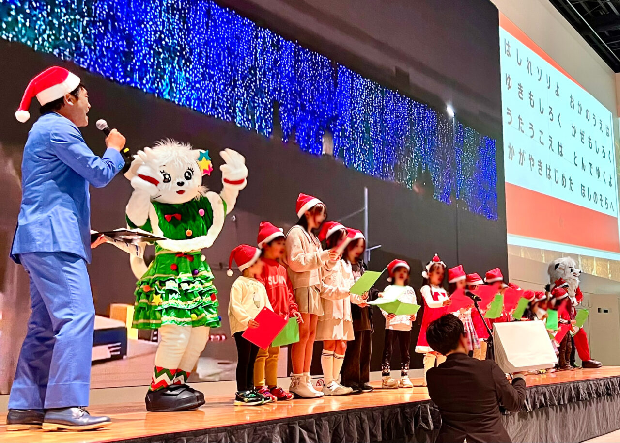 ライナツリー点灯式で生中継の壁面イルミネーションを背景に歌うサンタキッズたち