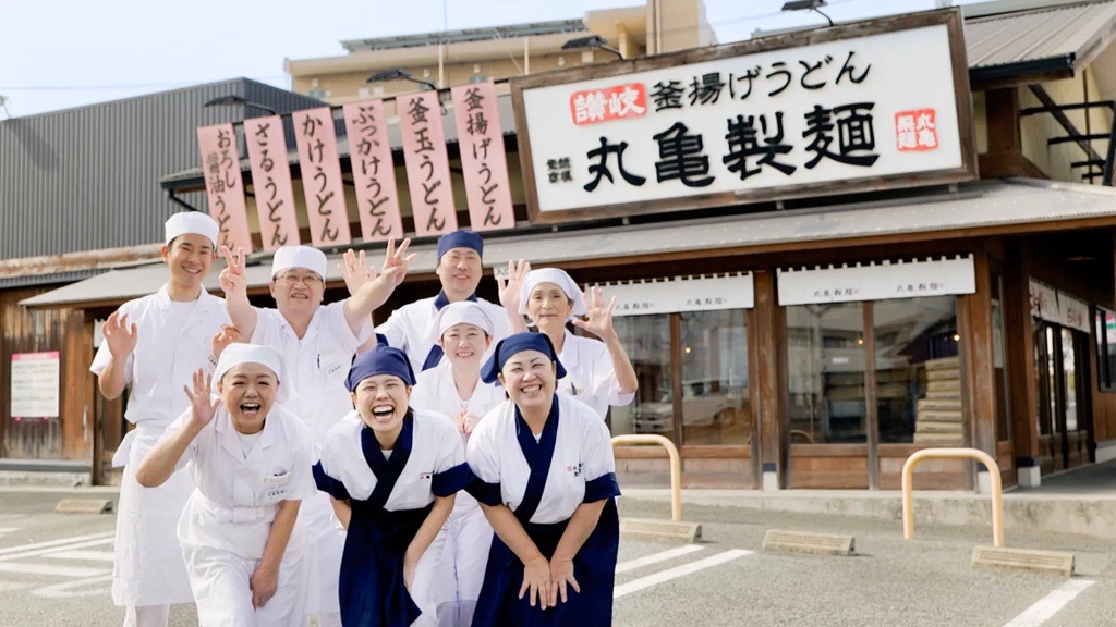 丸亀製麺の店舗前で笑顔のスタッフが並ぶ様子